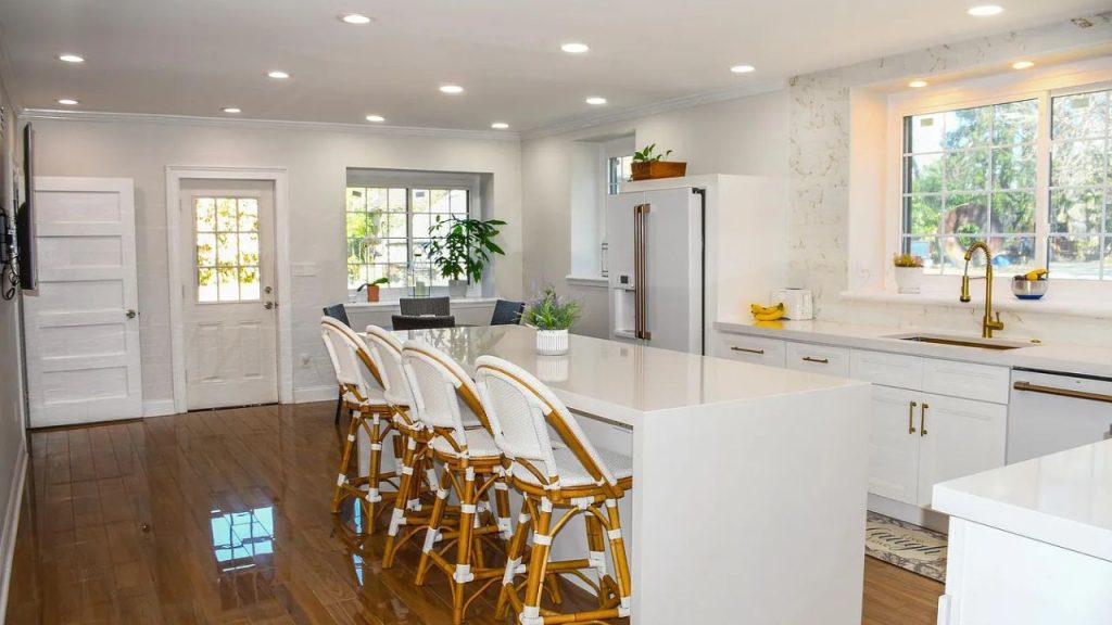 A bright kitchen with a white island, rattan stools, large windows, and potted plants on hardwood floors.
