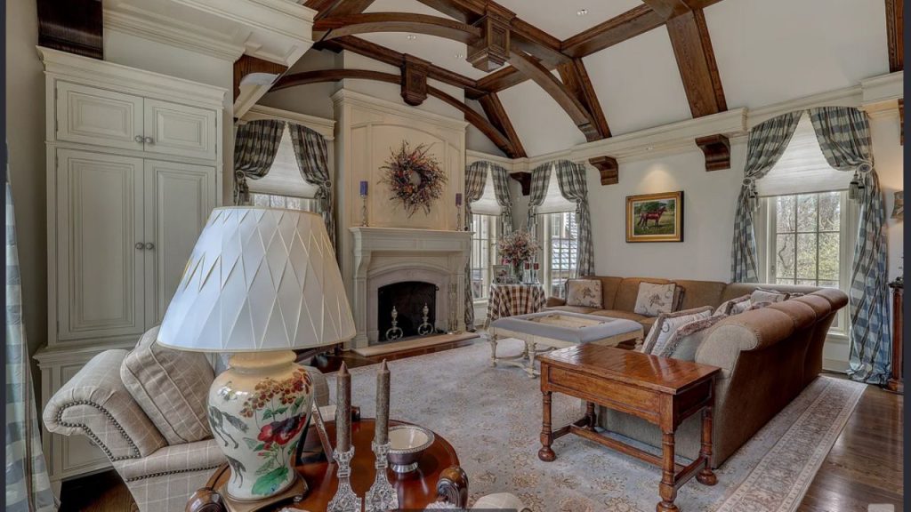 A cozy living room with wooden beams, a stone fireplace, plush sofas, and large windows with plaid curtains.