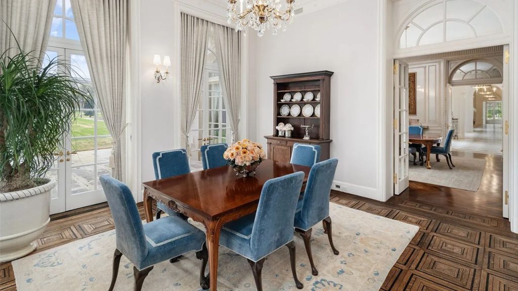 A cozy dining area with a wooden table, blue chairs, chandelier, and large windows.