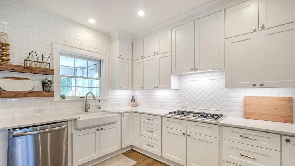 A bright kitchen with white cabinets, marble countertops, a farmhouse sink, and open shelving under large windows.