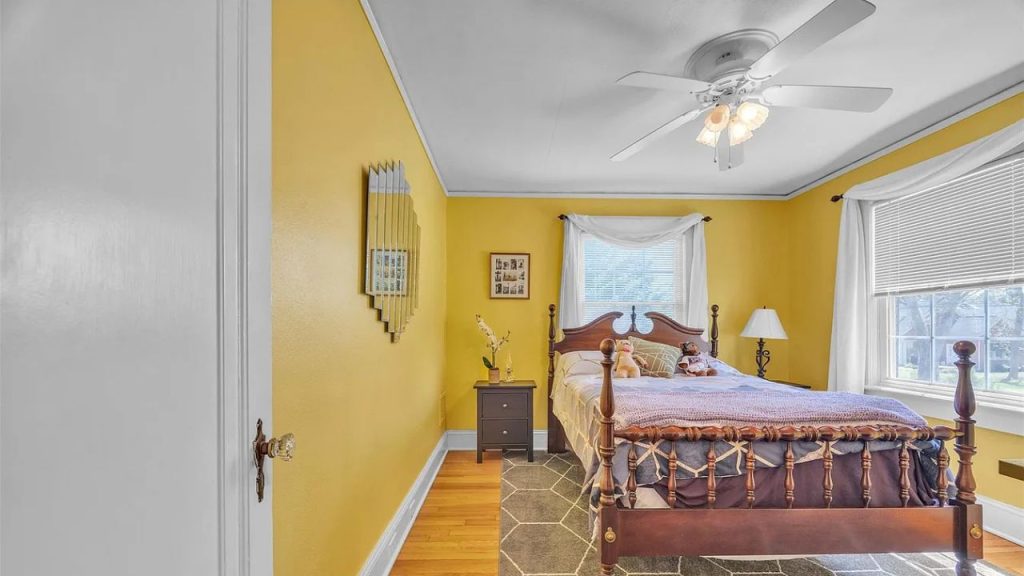 A cheerful bedroom with yellow walls, a wooden bed, white curtains, a ceiling fan, and a geometric rug.