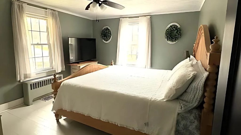 A peaceful bedroom with a wooden bed, white linens, a ceiling fan, and large windows with wreaths on the walls.