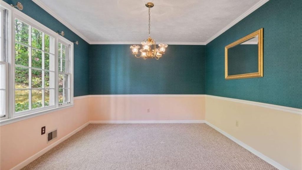 A stylish dining room with teal walls, peach wainscoting, a chandelier, and a large mirror, brightened by big windows.