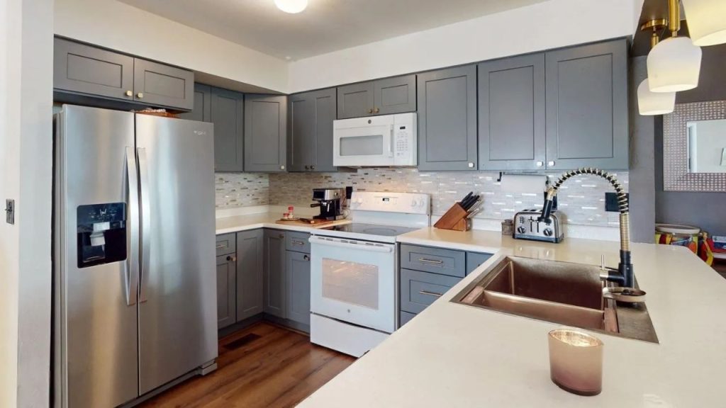 A modern kitchen with gray cabinets, stainless-steel fridge, white oven, and a sink with a pull-down faucet, lit by pendant lamps.