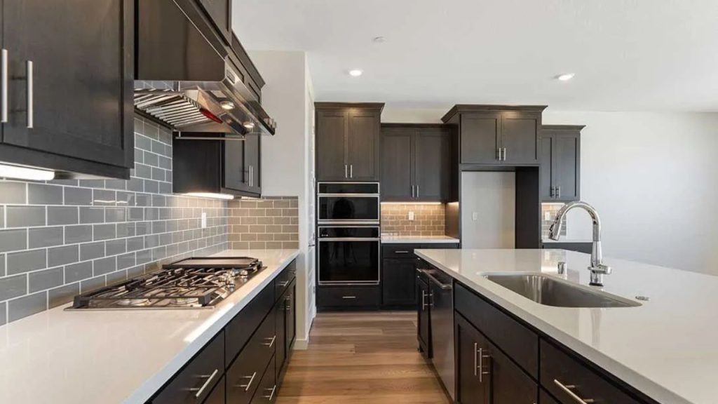 A modern kitchen with dark cabinets, white quartz countertops, a large island with a sink, stainless steel appliances, gray subway tile backsplash, and warm wood floors.