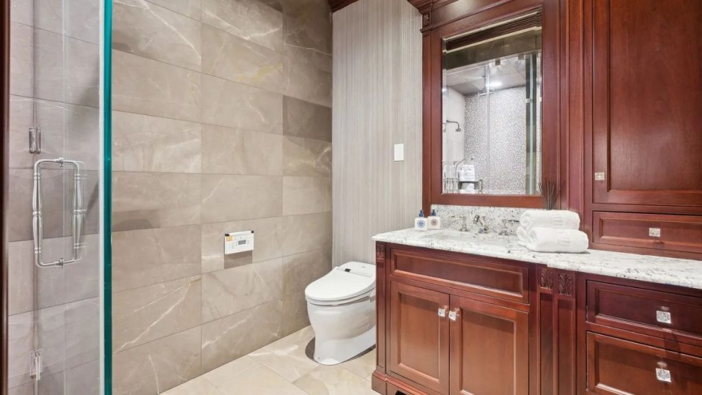 A guest bathroom with mahogany cabinets, a marble countertop vanity, a modern toilet, and a glass shower, featuring large veined tiles and soft lighting.