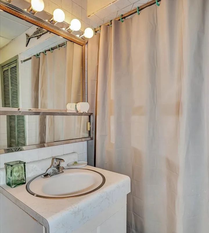 A vintage bathroom with white tiles, beige curtains, an oval sink, Hollywood-style vanity lights, and a fish sculpture on the wall, creating a serene atmosphere.
