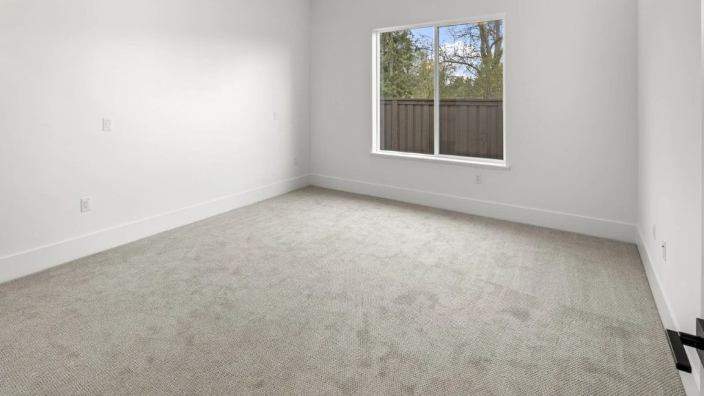 A minimalist bedroom with gray carpeting, white walls, and a large window overlooking a fenced backyard with trees.