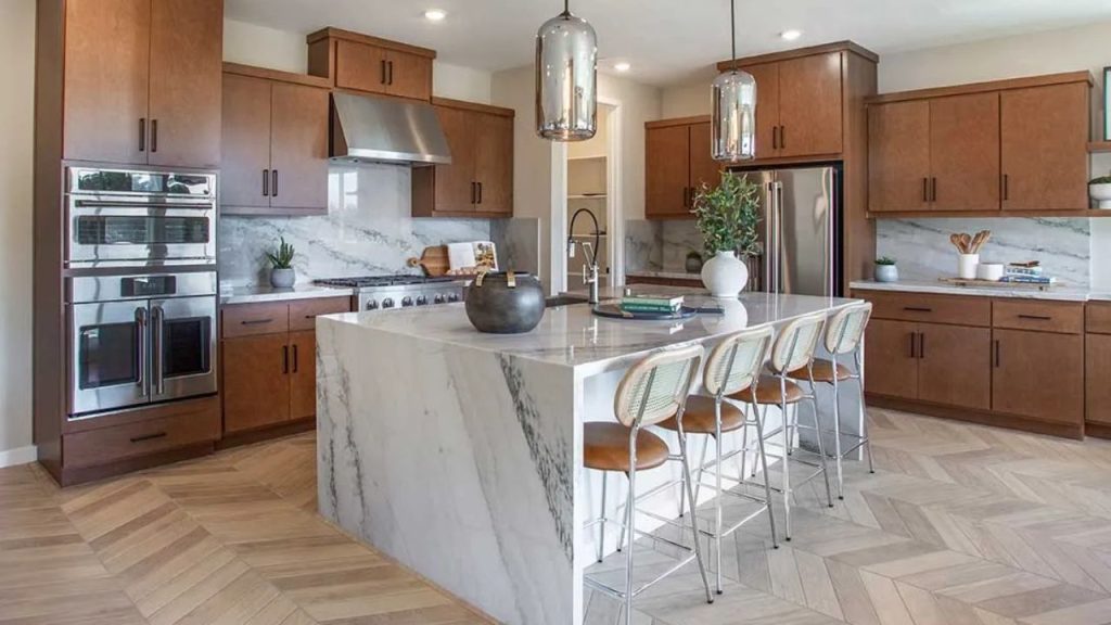 A spacious kitchen with warm wooden cabinets, a marble island, pendant lights, and a herringbone wood floor.