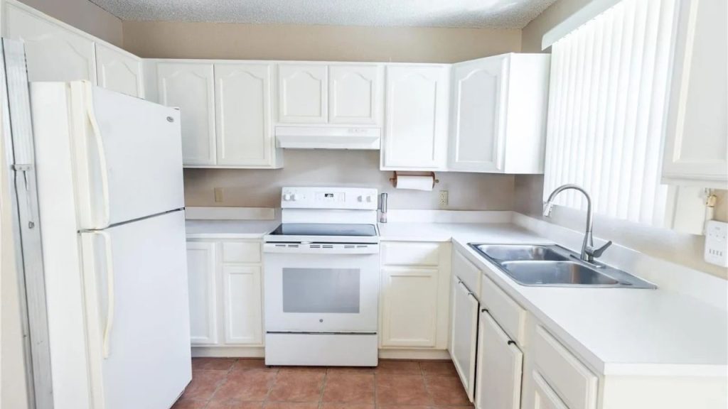 A bright kitchen with white cabinets, a double sink, electric stove, refrigerator, and terracotta tiles.
