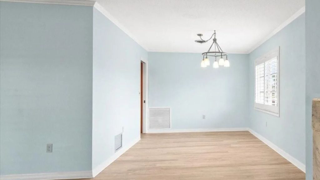 A bright dining area with pale blue walls, light wood floors, white shutters, and a chandelier with exposed bulbs over an open space.