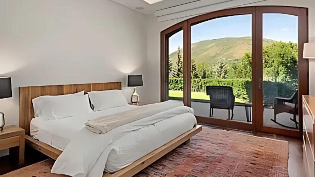 A cozy bedroom with a wooden bed, white linens, and arched doors opening to a mountain view, featuring a warm rug and balcony chairs.