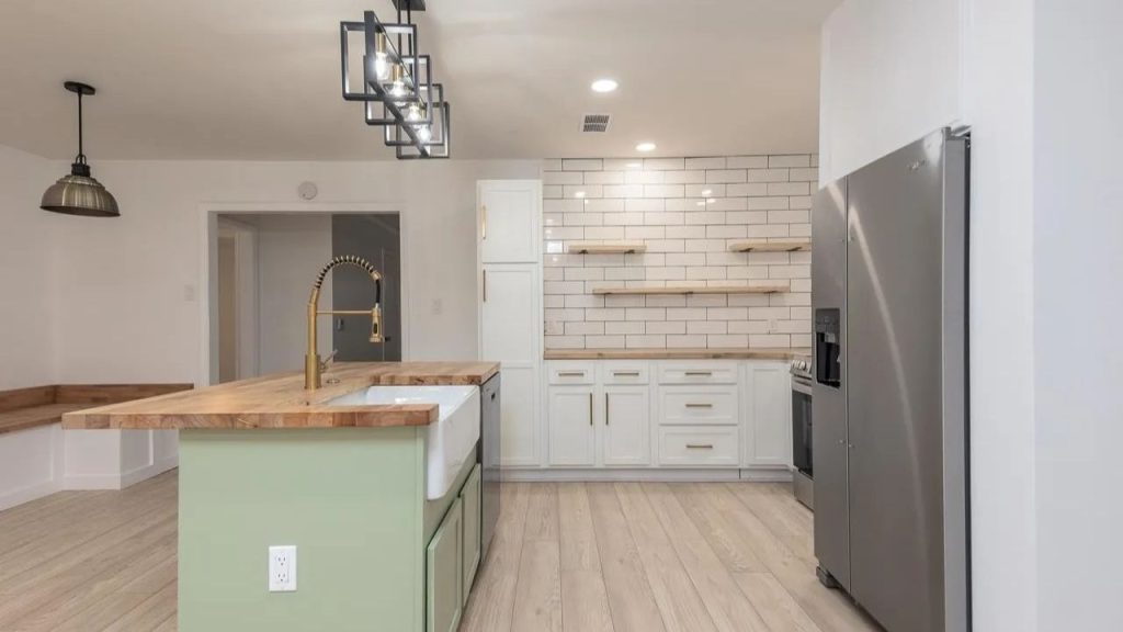 A rustic kitchen with a farmhouse sink, sage green island, and white subway tiles, accented by modern lights and a stainless steel fridge.