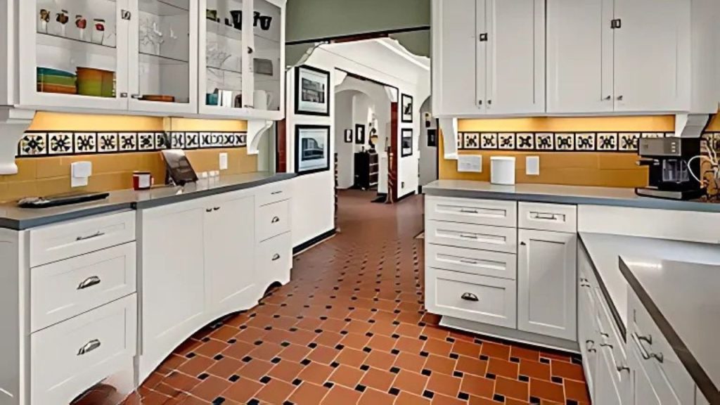 A bright kitchen with white cabinets, terracotta tile floor, and a decorative backsplash, opening to an adjacent room with artwork.