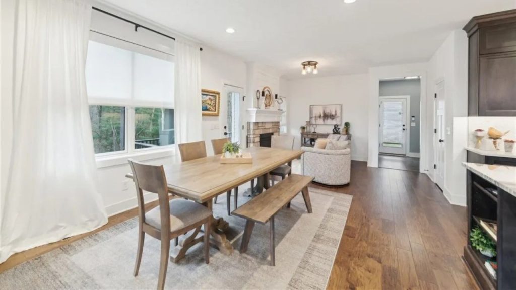 A cozy dining area with a wooden table, stone fireplace, and large windows with sheer curtains.