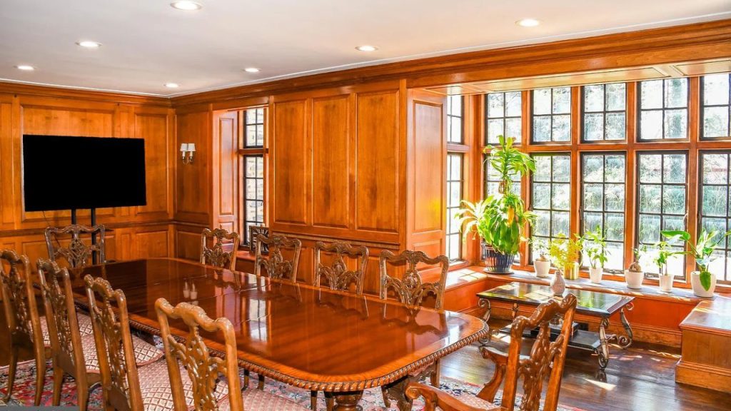 An elegant dining room with wooden paneling, a long polished table, large windows, and potted plants.