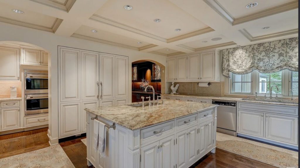 A bright kitchen with a large granite island, white cabinets, high coffered ceilings, and floral-curtained windows.