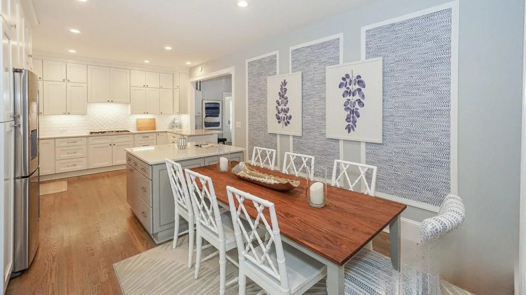 An elegant dining area with a wooden table, white chairs, textured walls, and a view of a modern kitchen.