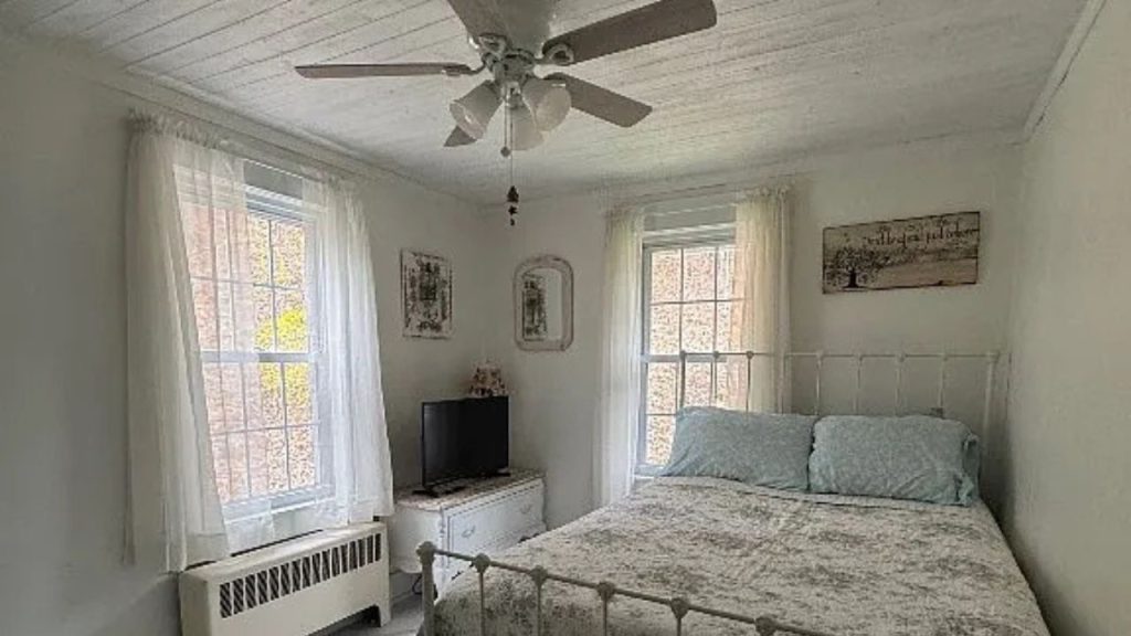 A quiet bedroom with an iron bed, gray quilt, ceiling fan, and large windows with sheer curtains.