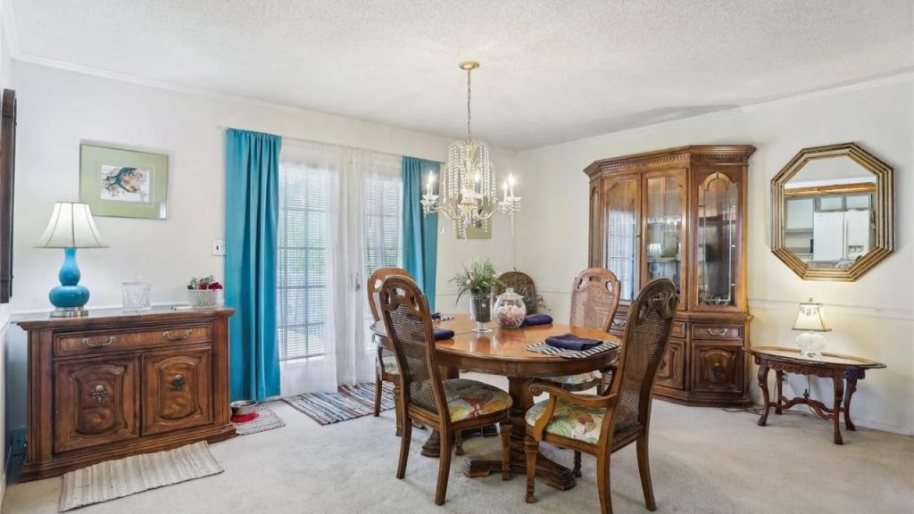 A cozy dining area with a wooden table, carved chairs, crystal chandelier, and blue curtains, featuring a hutch and mirror.