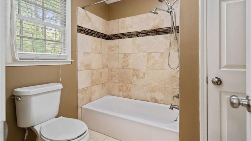 A calm bathroom with beige walls, a tiled shower-tub, a window with greenery, and a white toilet.