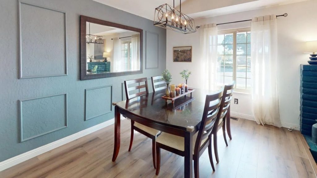 A cozy dining area with a wooden table, chandelier, teal wall, large mirror, and sheer-curtained windows.
