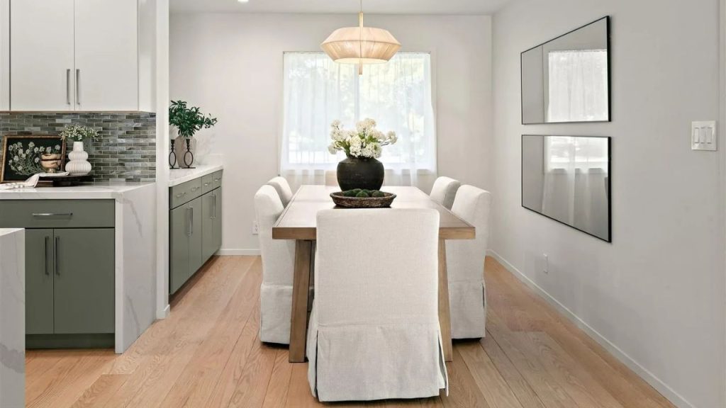 A bright dining area with a wooden table, upholstered chairs, and mirrors, next to a stylish kitchen.