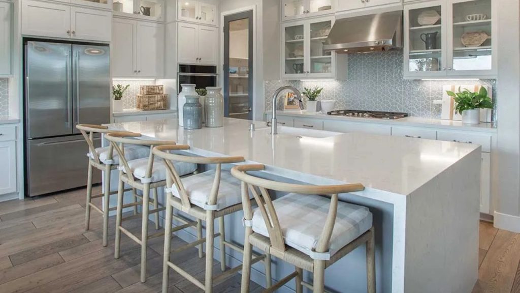 A bright kitchen with white cabinets, a gray geometric backsplash, a large island with woven barstools, stainless steel appliances, and light wood floors, accented by plants and rustic decor.