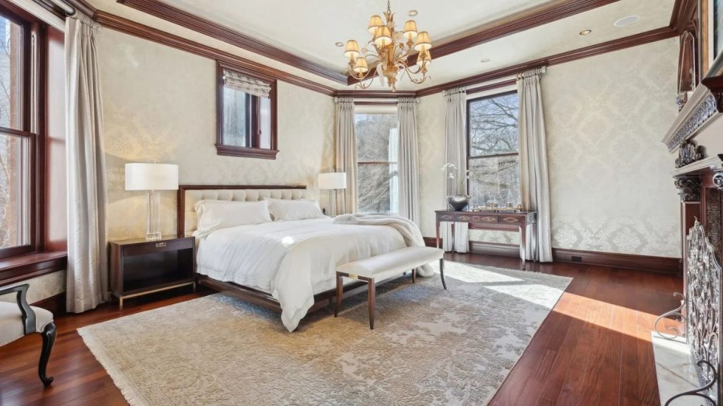 A serene bedroom with a plush bed, tufted headboard, chandelier, and fireplace, featuring tall windows with cream curtains and hardwood floors.