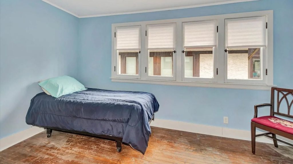 A serene bedroom with pale blue walls, a bed with a navy quilt, large windows with white shades, hardwood floors, and a wooden chair with a red cushion in the corner.