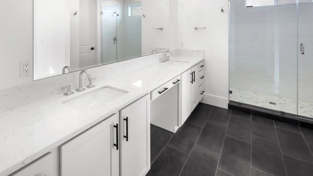 A modern primary bathroom with a double vanity, marble countertop, dark slate floor tiles, a glass shower with subway tiles, and a large mirror.