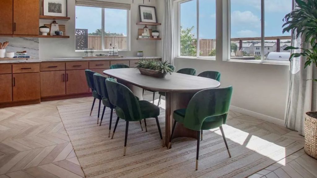 A bright dining area with a wooden table, green velvet chairs, large windows, and a herringbone floor.
