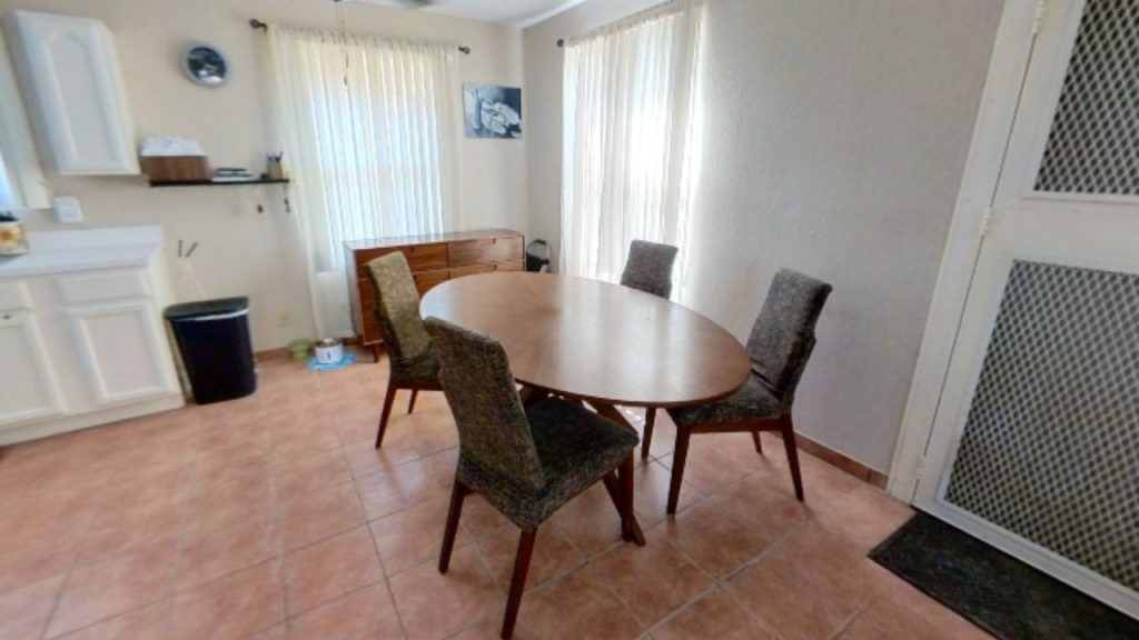 A cozy dining area with a round wooden table, four chairs, wooden cabinet, and sunlight through sheer curtains.