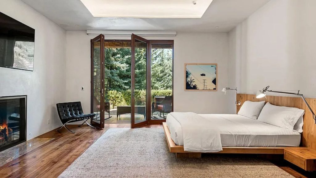 A cozy bedroom with a wooden bed, white linens, folding glass doors to a green view, a fireplace, and a black chair.