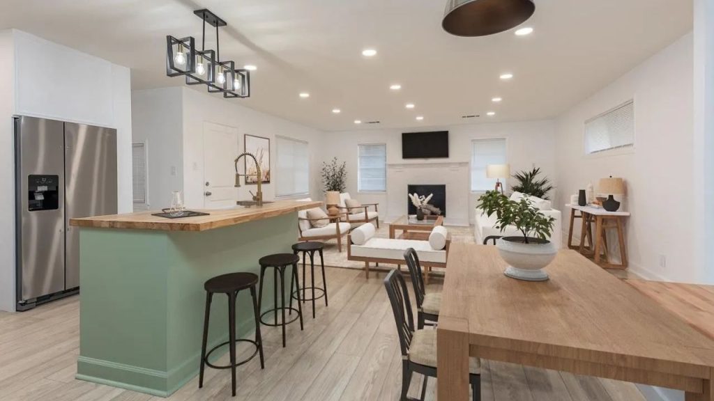 An open-plan kitchen with a sage-green island, wooden top, and bar stools, flowing into a living area with a fireplace and dining table.