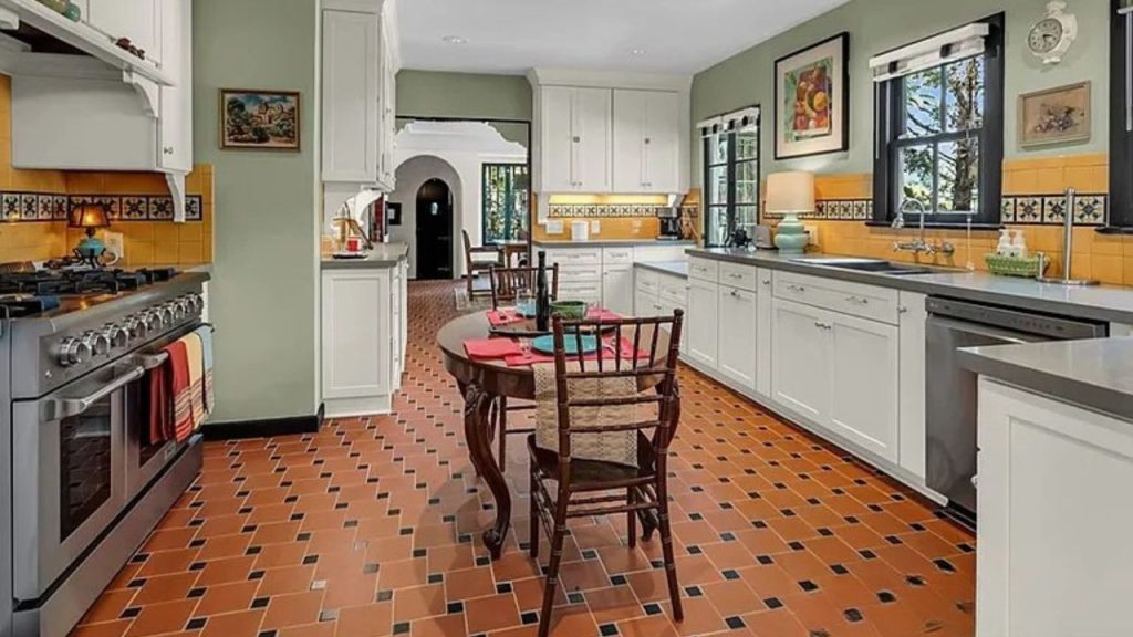 A cozy kitchen with terracotta tile floor, white cabinets, a yellow backsplash, and a dining table by large windows.
