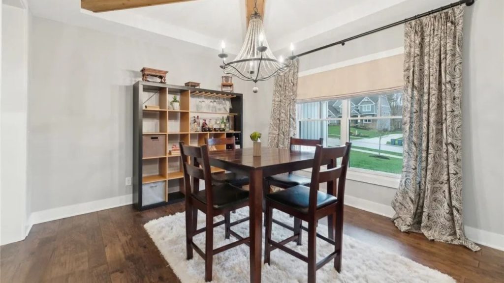 A cozy dining nook with a wooden table, chandelier, and large windows overlooking a green lawn.