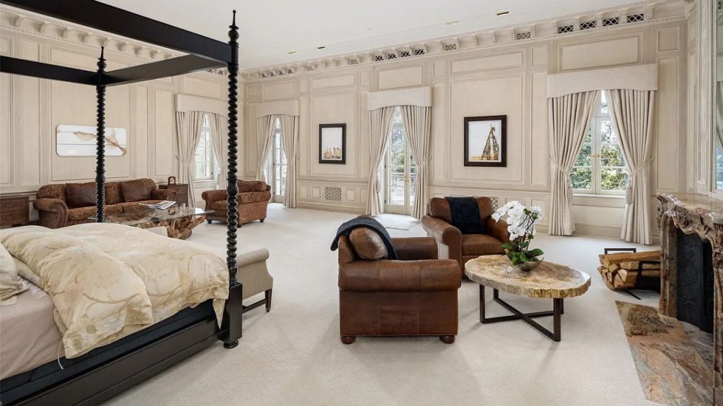 A luxurious bedroom with a four-poster bed, seating areas, a fireplace, and large windows with draped curtains.