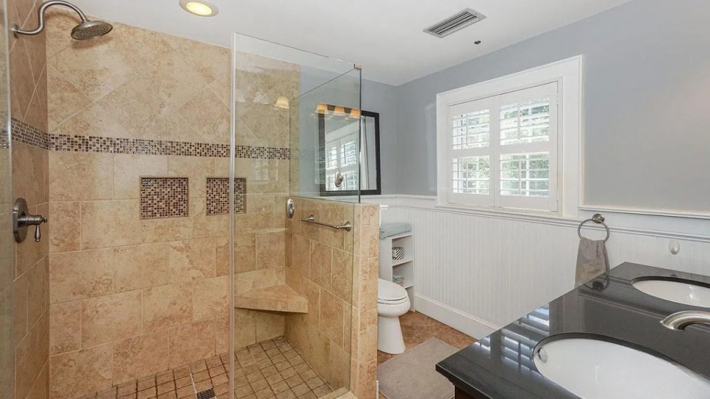 A spacious bathroom with beige tiled shower, dual sinks, large window, and built-in shelves.
