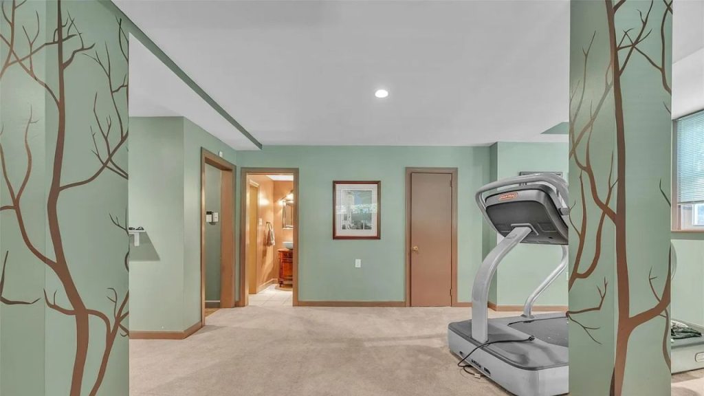 A cozy home gym with a treadmill, green walls with tree designs, carpeted floor, and a glimpse of an adjacent bathroom.