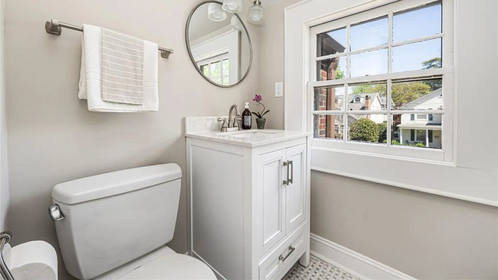 A bright bathroom with a white vanity, round mirror, and large window.