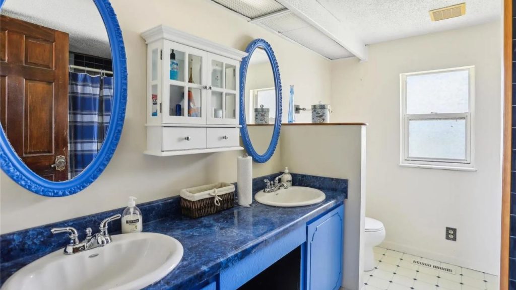 A bright bathroom with blue countertops, double sinks, and mirrors, featuring a white cabinet and tiled floor under soft natural light.