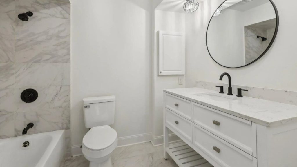 A bright bathroom with a bathtub, glass shower, white vanity, and round mirror, featuring marble tiles.