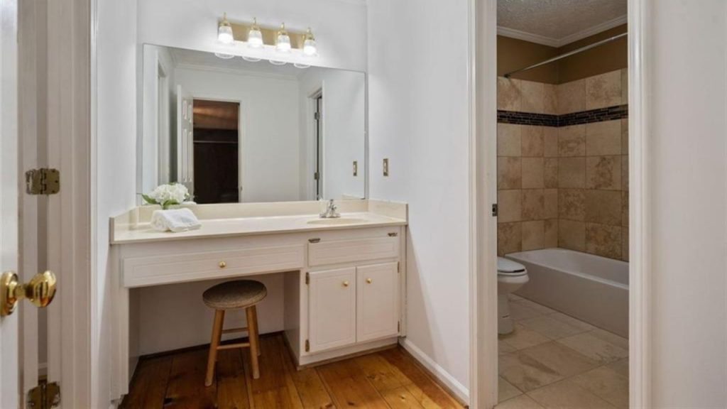 A tranquil vanity area with a mirror, white cabinetry, wooden stool, and a glimpse of a tiled bathroom with a tub.