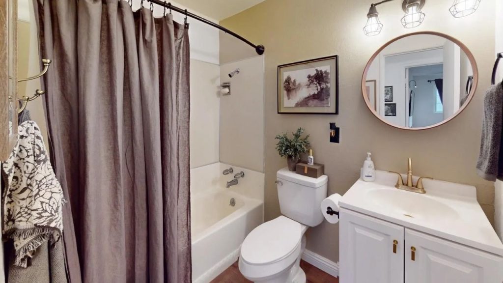A cozy bathroom with a bathtub, white sink, golden faucet, round mirror, and soft lighting, featuring a gray curtain and plant.
