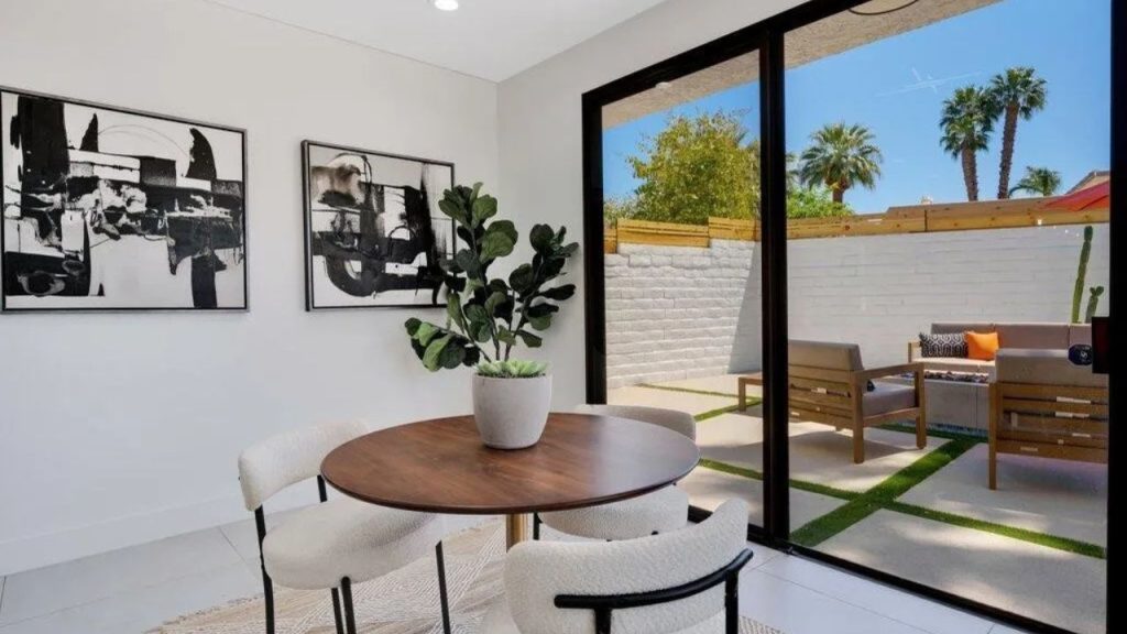 A bright dining nook with a wooden table, white chairs, and views of a patio with palm trees.