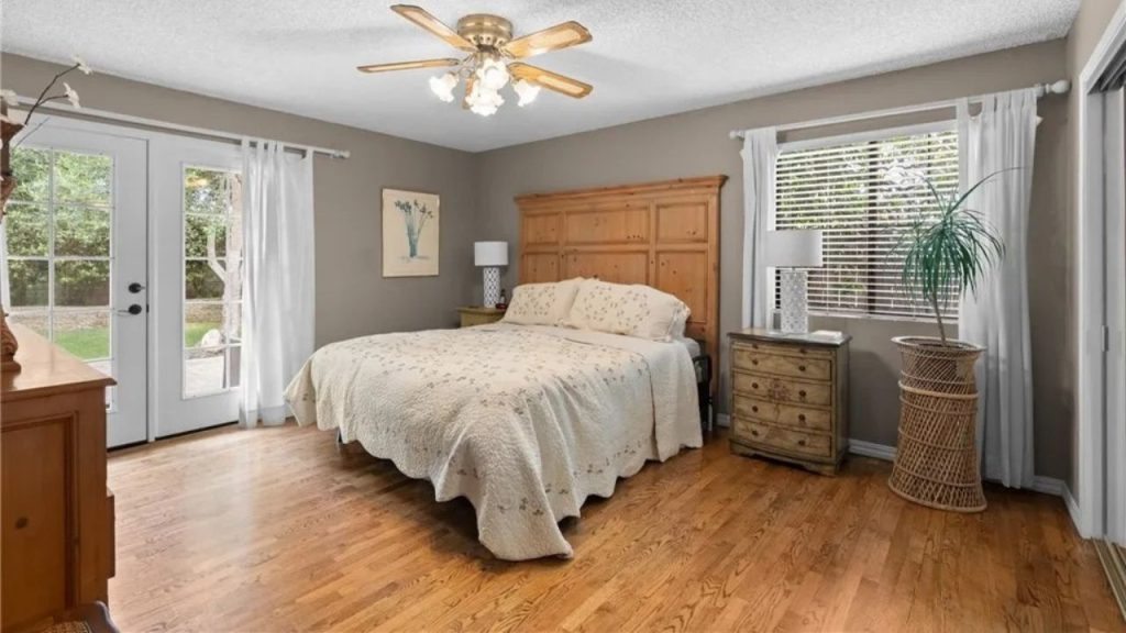 A relaxing bedroom with a wooden bed, hardwood floors, large windows, and wicker plant decor.