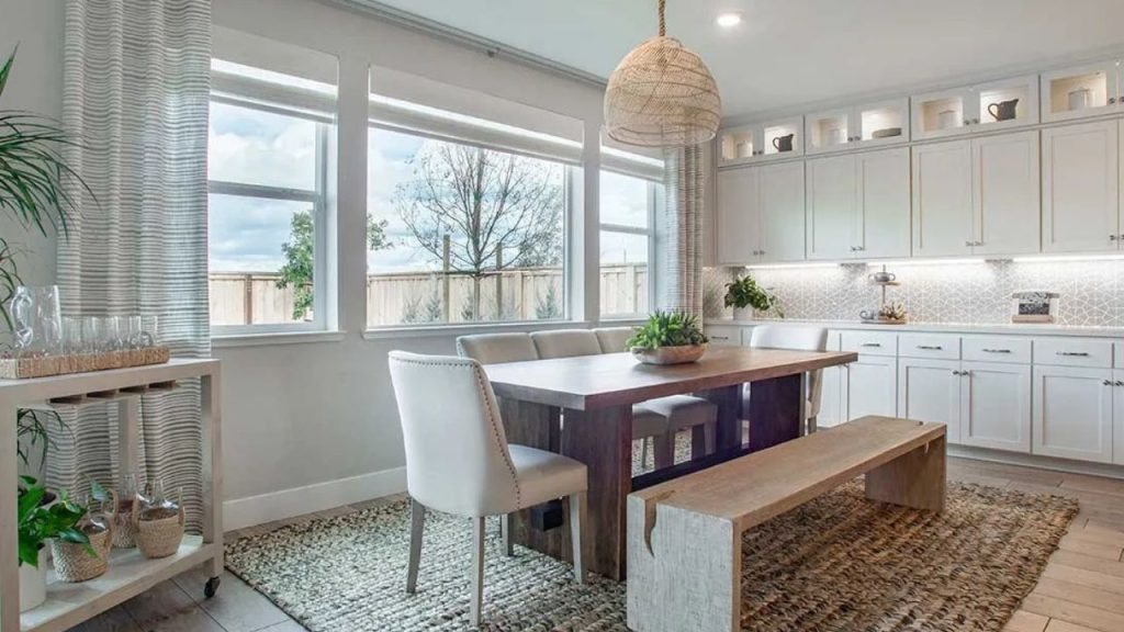 A cozy dining area with a wooden table, cushioned chairs, a bench, large windows with sheer curtains, a woven pendant light, and light wood floors, adjacent to a kitchen with white cabinets.