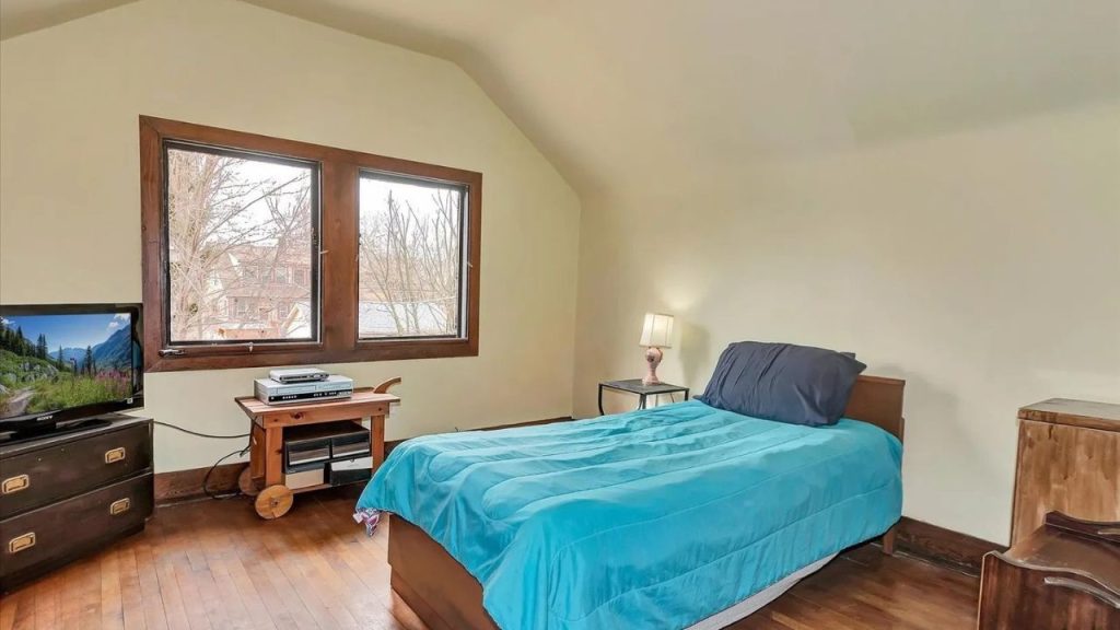 A minimalist attic bedroom with beige walls, a turquoise quilted bed, large windows, hardwood floors, a small nightstand with a lamp, and a vintage TV cart.