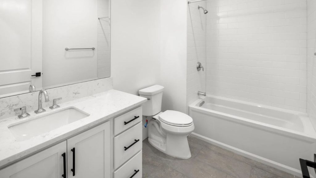 A guest bathroom with a white vanity, marble countertop, soaking tub, subway tile walls, light gray flooring, and a large mirror.